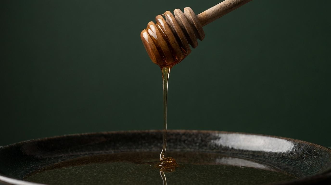 Amber honey flowing from a wooden dipper into a dark bowl against a forest-green background
